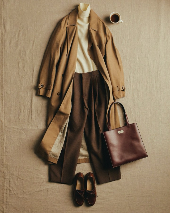 Fall outfit laid out on linen: a camel trench coat, cream turtleneck, brown trousers, suede loafers, and a leather tote.
